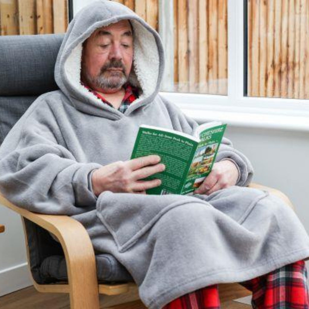 Man sitting in a chair wearing a grey hooded blanket, reading a green book.